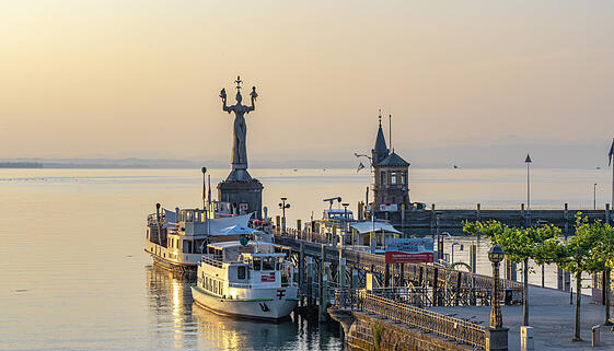 Blick auf die Hafeneinfahrt mit Imperia-Statue, dem Bodensee mit Katamaran F&auml;hre und Landschaftspanorama bei Sonnenaufgang. Konstanz, Bodensee, Baden-W&uuml;rttemberg, Deutschland, Europa.
