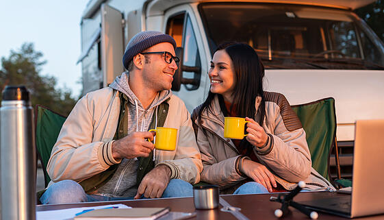 Smiling man holding cup with tea and squints against the bright sun near his woman. Happy family sitting at the nature and drinking hot tea from cups at camp fire in cozy forest