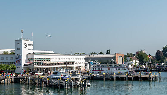 Der Hafen der Stadt Friedrichshafen am Bodensee mit Schiffen und einem Zeppelin im Hintergrund Der Hafen der Stadt Friedrichshafen am Bodensee mit Schiffen und einem Zeppelin im Hintergrund
