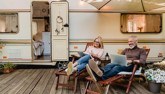Pleased happy aged couple reading a book and using laptop while relaxing on the deck chairs near the camper van